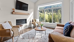 A living room with a fireplace, a large window with a forest view, a flat-screen TV, and a cozy seating area. - Modern & Cozy Condo w/Hot Tub! (Sun Peaks)