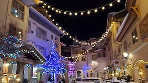 A snow-covered street with buildings on either side, decorated with string lights and illuminated trees. - Modern & Cozy Condo w/Hot Tub! (Sun Peaks)