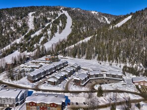 Un centro de esquí cubierto de nieve con varios edificios, incluyendo un hotel, un área de estacionamiento y un telesilla visible al fondo.