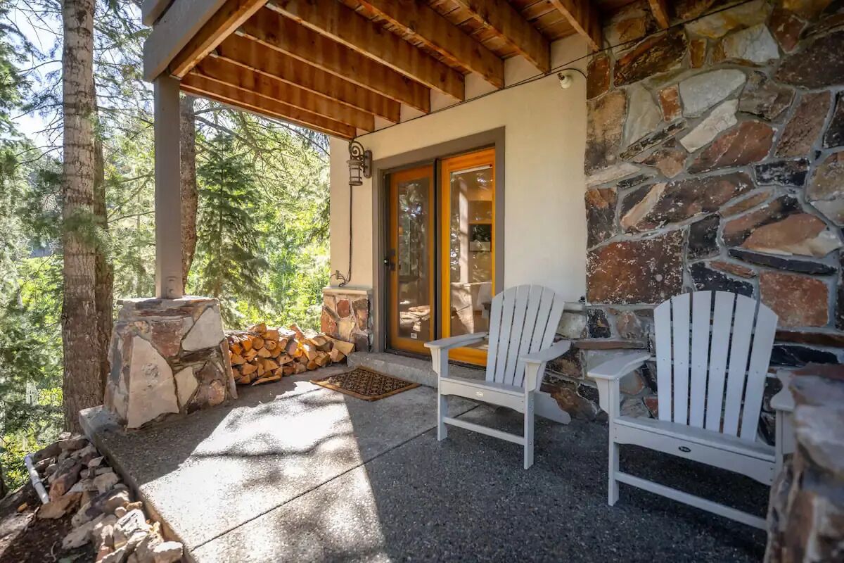 A covered outdoor seating area with two white Adirondack chairs, a stone fireplace, and a stone wall.