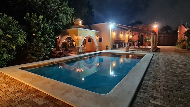 A swimming pool with a tiled deck, a covered seating area with chairs, and a building with arched doorways and outdoor lighting. - Espectacular casona con alberca y jardines (Mérida)