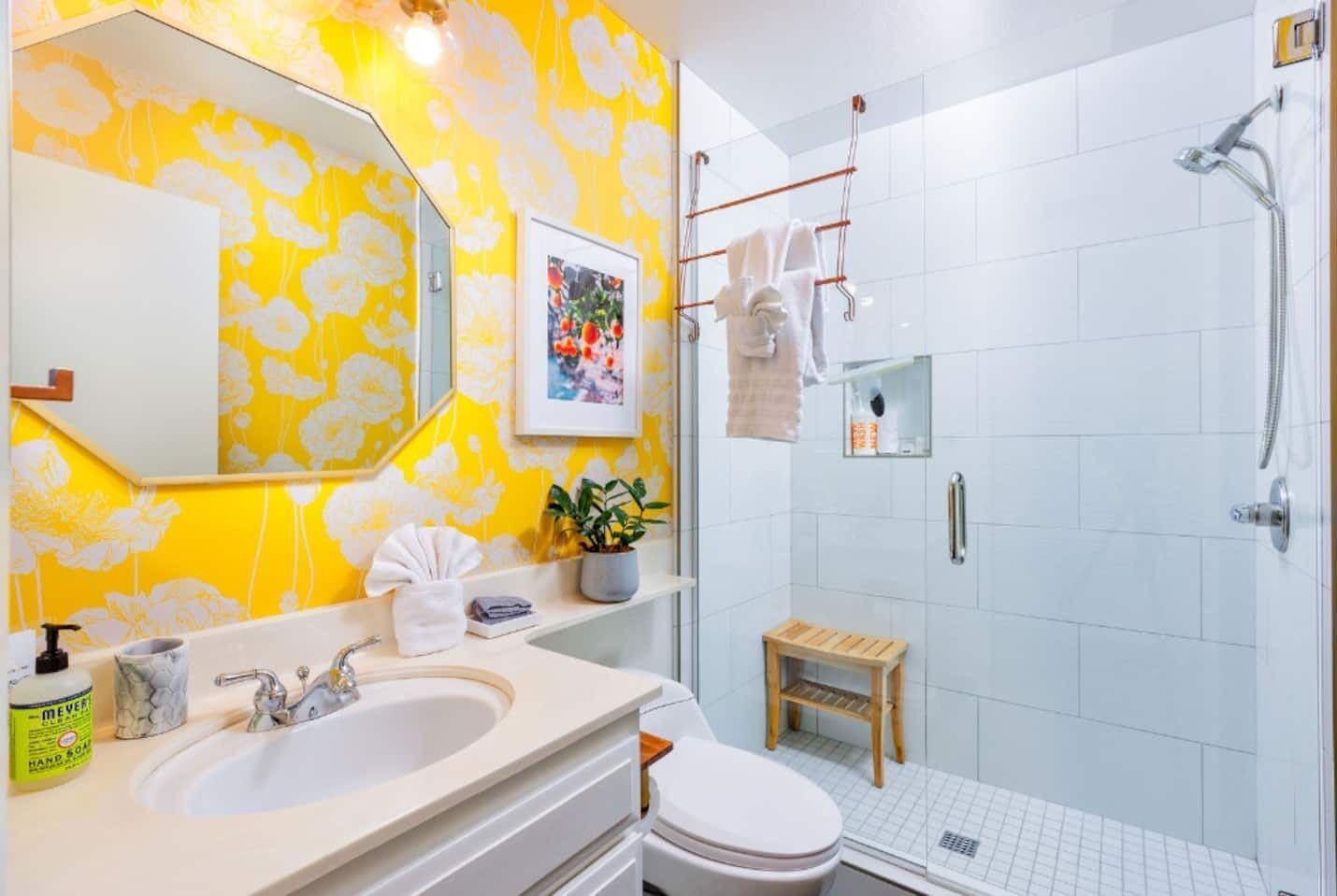 Ein Badezimmer mit gelbem floralem Wandpapier, einem weißen Waschbecken, einer Toilette, einer Dusche mit Glastür und einem kleinen Holzschemel.