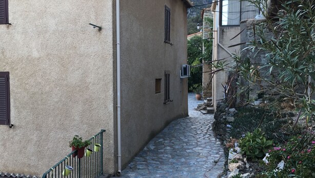 Une ruelle étroite pavée, bordée de bâtiments aux murs de pierre et aux volets fermés, qui mène vers un paysage montagneux en arrière-plan.