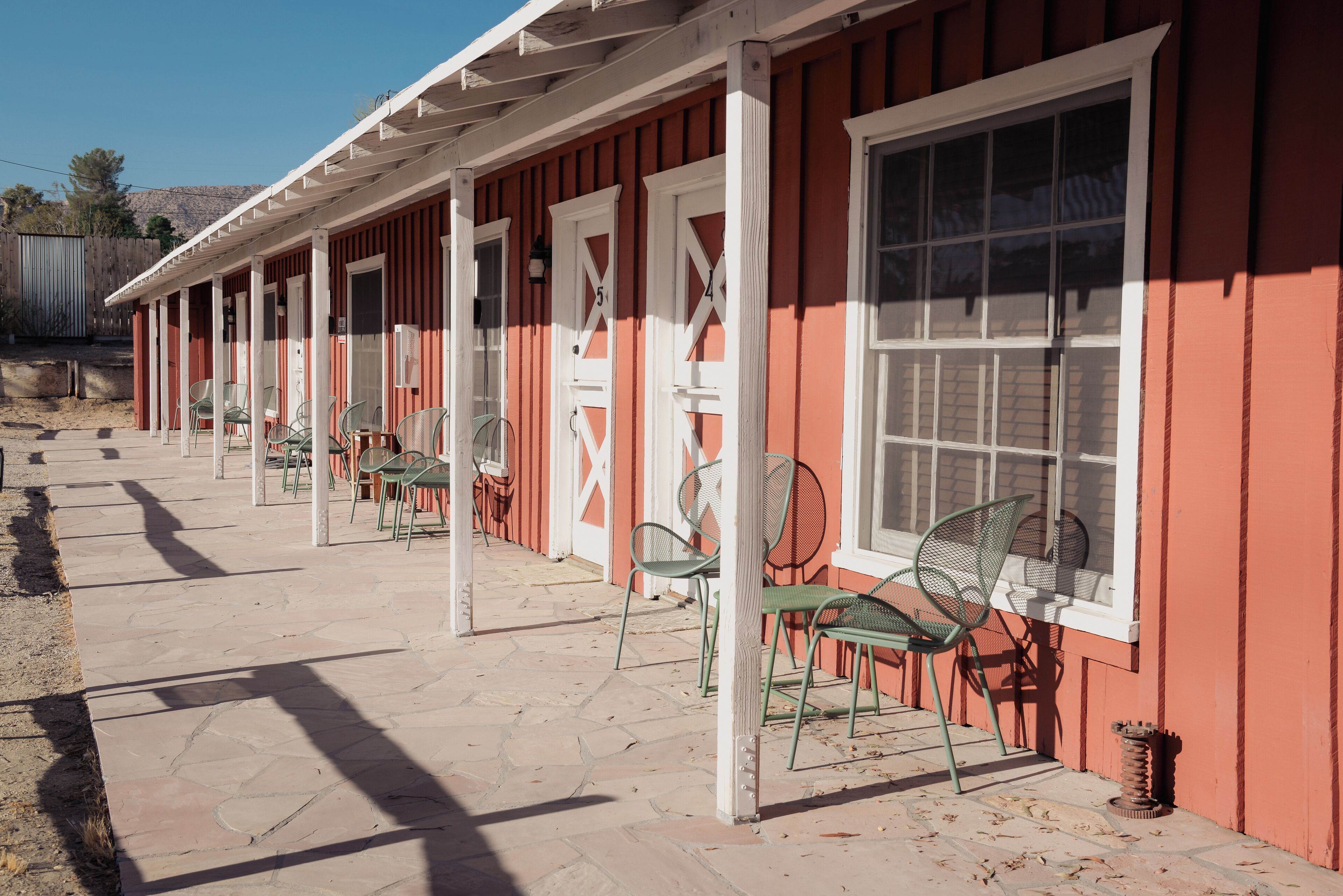 A row of red buildings with white-framed windows and doors, outdoor seating with green chairs, and a stone-paved walkway.