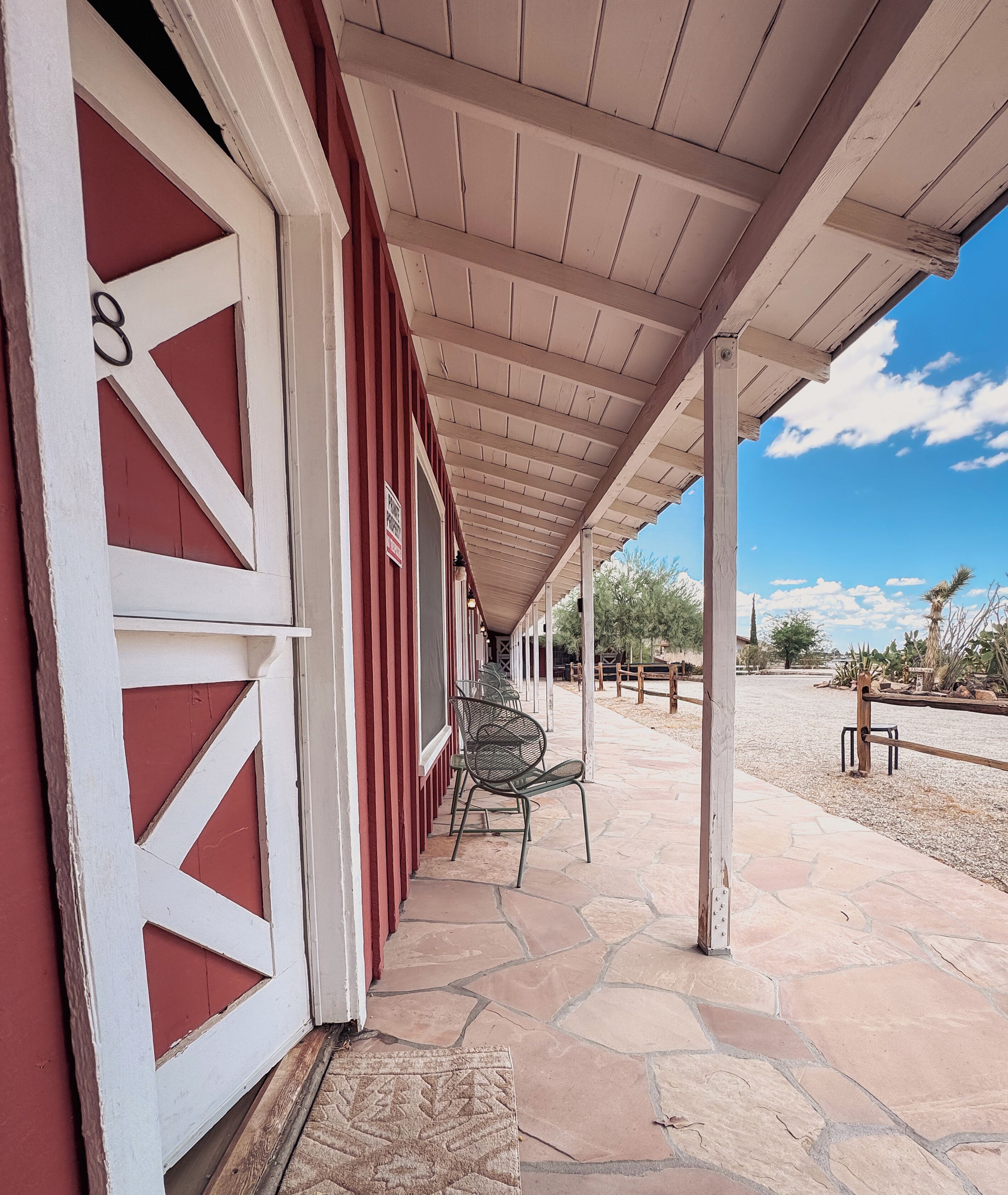 A covered outdoor area with a red building, stone flooring, and metal chairs.