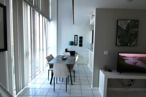 A modern dining area with a table and chairs, a TV on a stand, and a window with blinds. - Castle B6  Bayview View Loft, Pool and Beach Access, Gym and Free Valet Parking (Miami Beach)