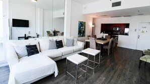 Una sala de estar moderna con un sofá blanco, dos mesas blancas y una cocina con armarios de madera y un microondas.