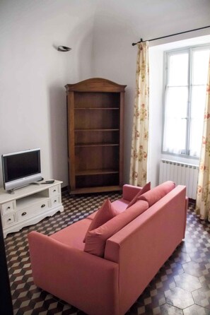 A room with a pink sofa, a television on a white cabinet, a wooden bookshelf, and a window with floral curtains. - Holiday apartment in the medieval village of Finale Ligure (Finale Ligure)