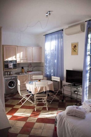 Ein kleines Zimmer mit Küchenzeile, einem Essbereich mit Tisch und Stühlen, einem Bett mit weißer Bettdecke und einem Fernseher auf einem Schrank.