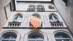 Eine Gebäudefassade mit drei Rundbogenfenstern und einem Balkon mit Pflanzen. Ein gelb-rot gestreifter Banner hängt zwischen den Fenstern.