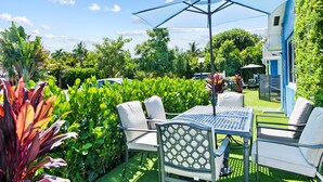 An outdoor patio with a table and chairs, surrounded by lush greenery and a clear blue sky.
