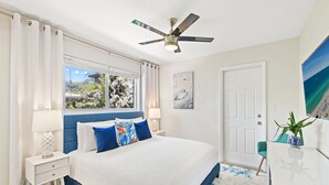 A bedroom with a large bed, a ceiling fan, a window with a view of trees, a door, and a painting on the wall.