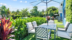 An outdoor patio with a table and chairs, surrounded by lush greenery and a clear blue sky.