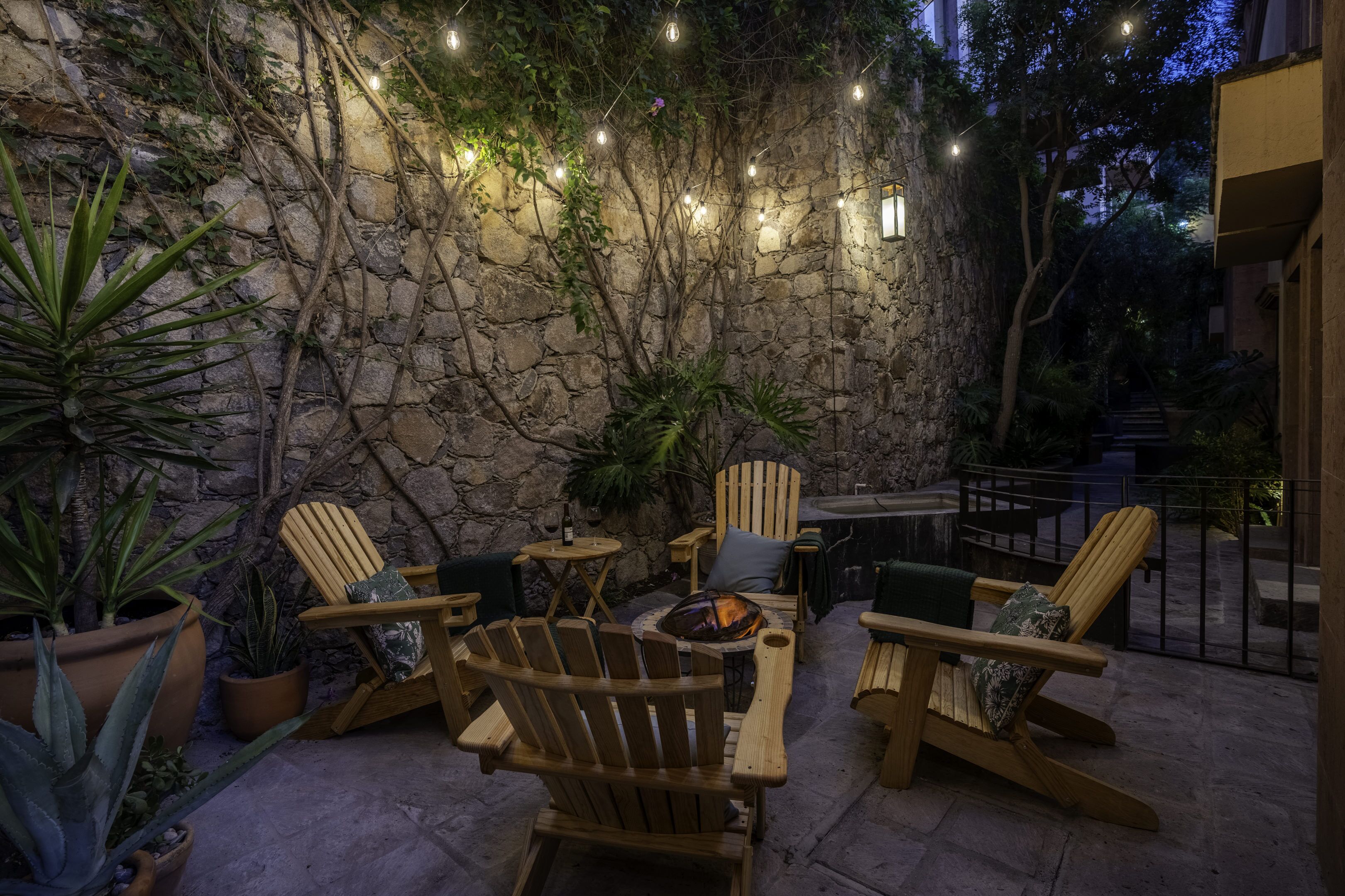 Un muro de piedra con luces colgantes, sillas de madera y un hogar en un patio al aire libre.