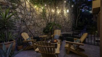 Un muro de piedra con luces colgantes, sillas de madera y un hogar en un patio al aire libre.