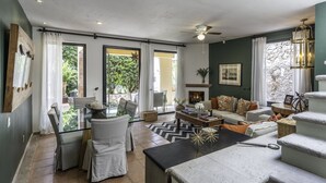 A spacious living room with a sofa, chairs, a coffee table, and a fireplace. There are large windows with curtains, a ceiling fan, and a stone wall. - Angelic Centro 2BR with Pool (San Miguel de Allende)