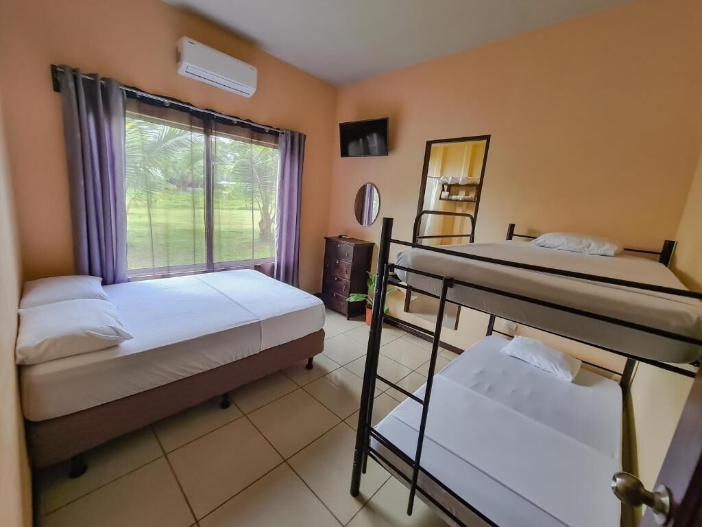 A room with two beds, a window with curtains, an air conditioning unit, a small table, and a mirror.