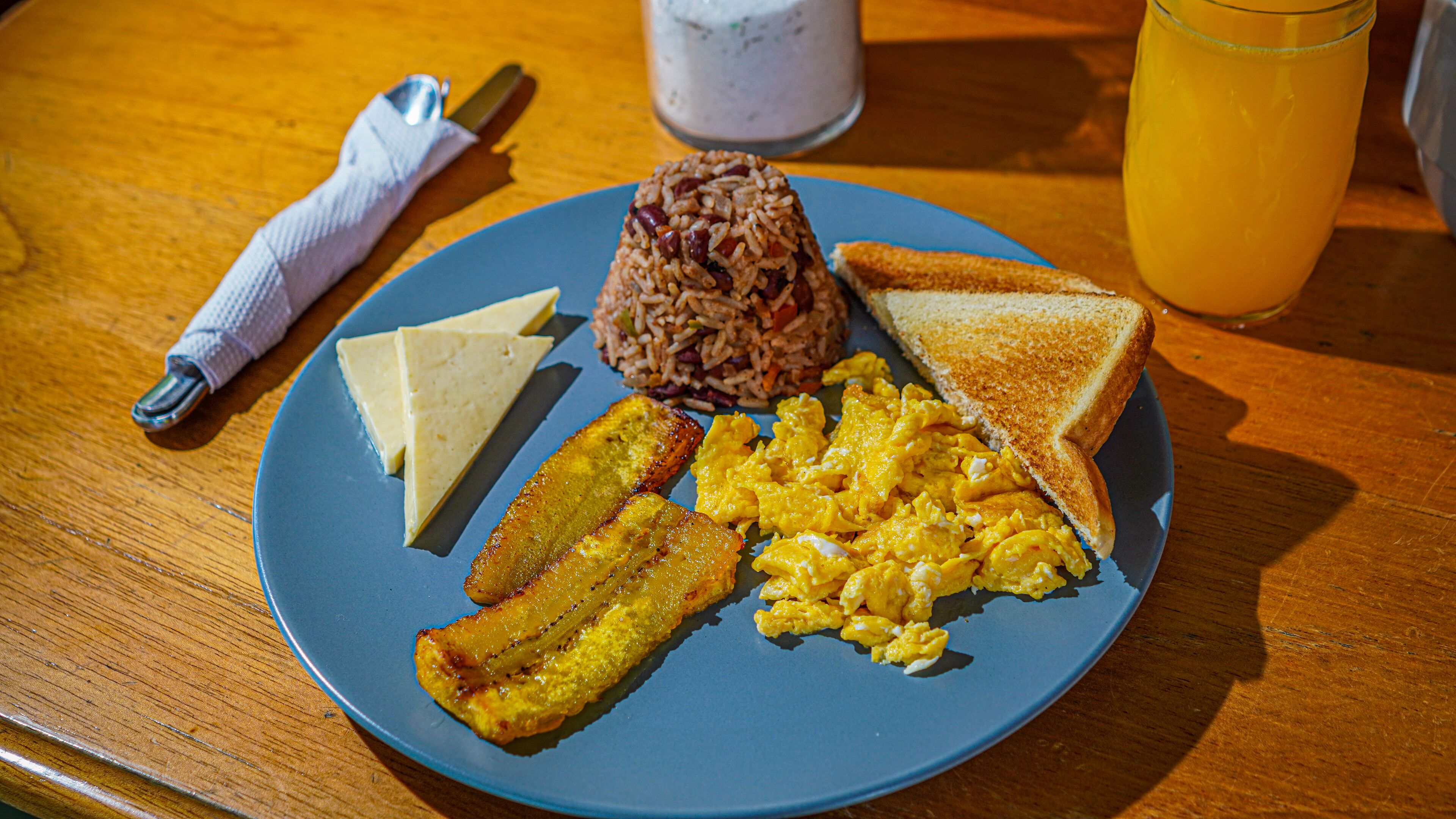 Un plato con huevos revueltos, plátanos fritos, arroz, tostadas y queso sobre una mesa de madera.