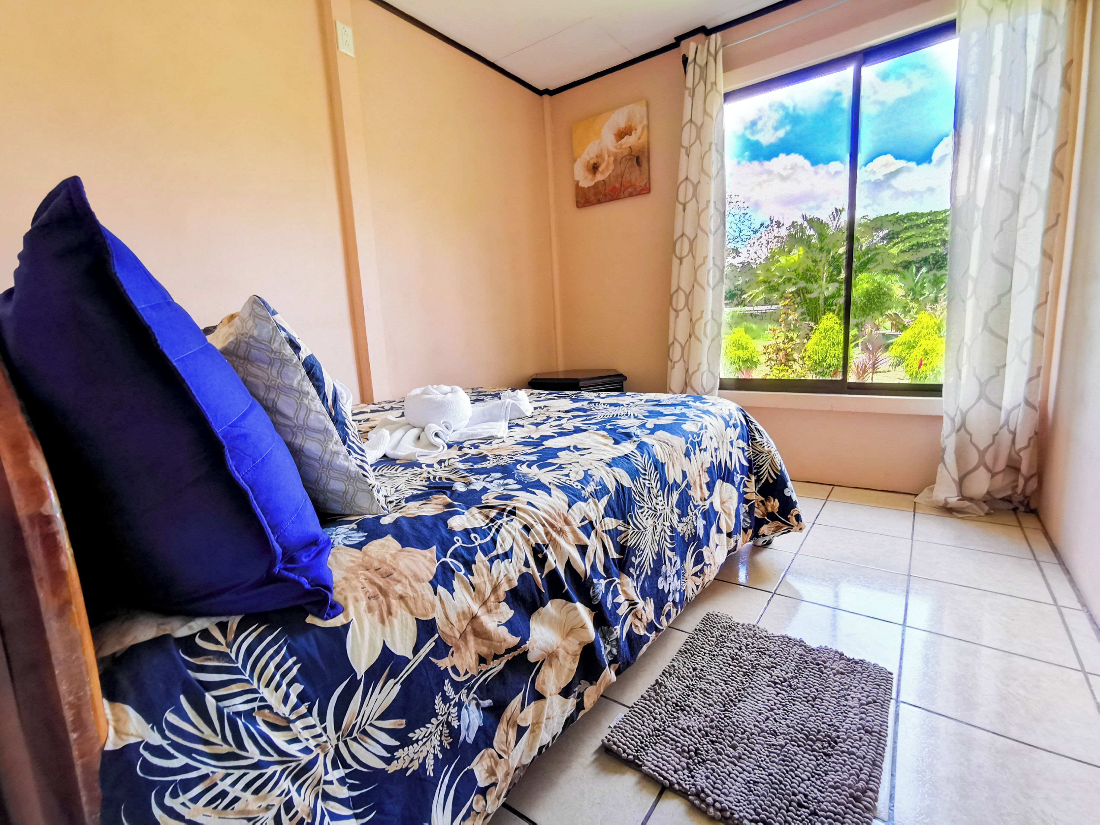 A bedroom with a bed, pillows, a bedside table, a window with a view, and a rug.