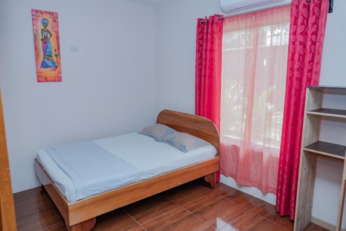 A single bed with a wooden frame, a white wall, a red curtain, and a small shelf unit.