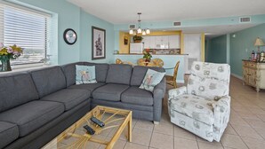 Una sala de estar con un sofá seccional gris, una mesita redonda con dos mandos a distancia, un sillón estampado y una zona de comedor con una cocina amarilla al fondo.