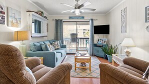 Ein Wohnzimmer mit einer Couch, zwei Sesseln, einem Couchtisch und einem Fernseher. Es gibt einen Balkon mit Blick auf Gebäude.