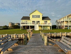 A two-story waterfront property with a wooden dock leading to a grassy lawn.