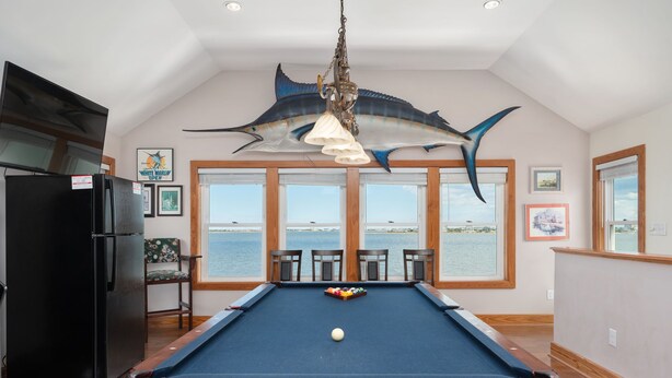 A pool table with a view of the sea through large windows, a black refrigerator, and a ceiling-mounted decoration. - A Speckled Trout: Soundfront, Hot Tub, Game Room, Private Dock (Nags Head)