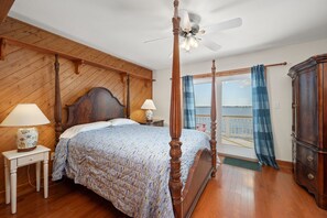A bedroom with a four-poster bed, wooden flooring, a ceiling fan, and a view of the sea through a balcony. - A Speckled Trout: Soundfront, Hot Tub, Game Room, Private Dock (Nags Head)