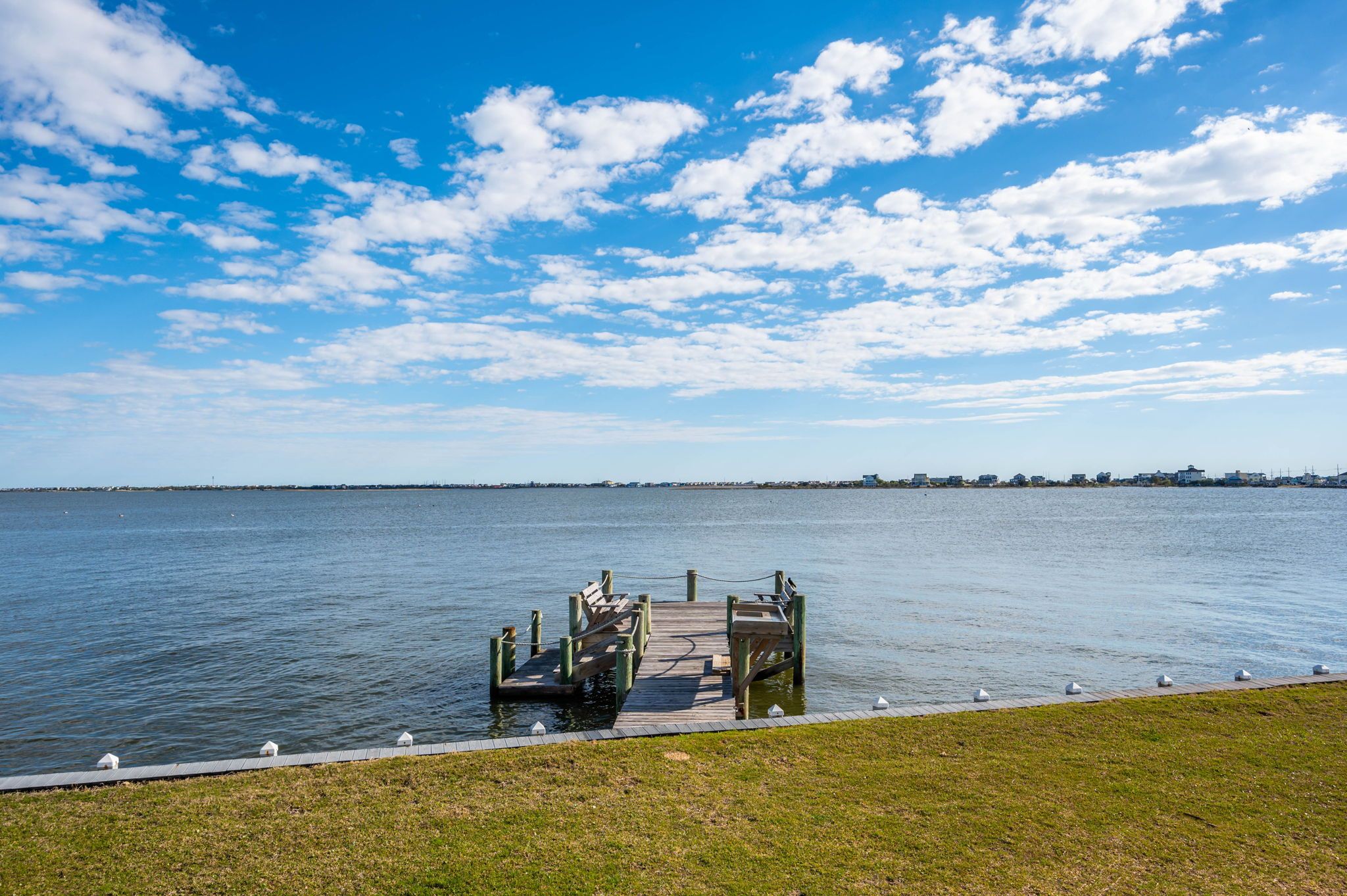 A wooden pier extending into calm water with a grassy area in the foreground and a clear sky above.