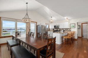 A spacious kitchen with a large island, wooden dining table, and chairs, with a view of a body of water through large windows.