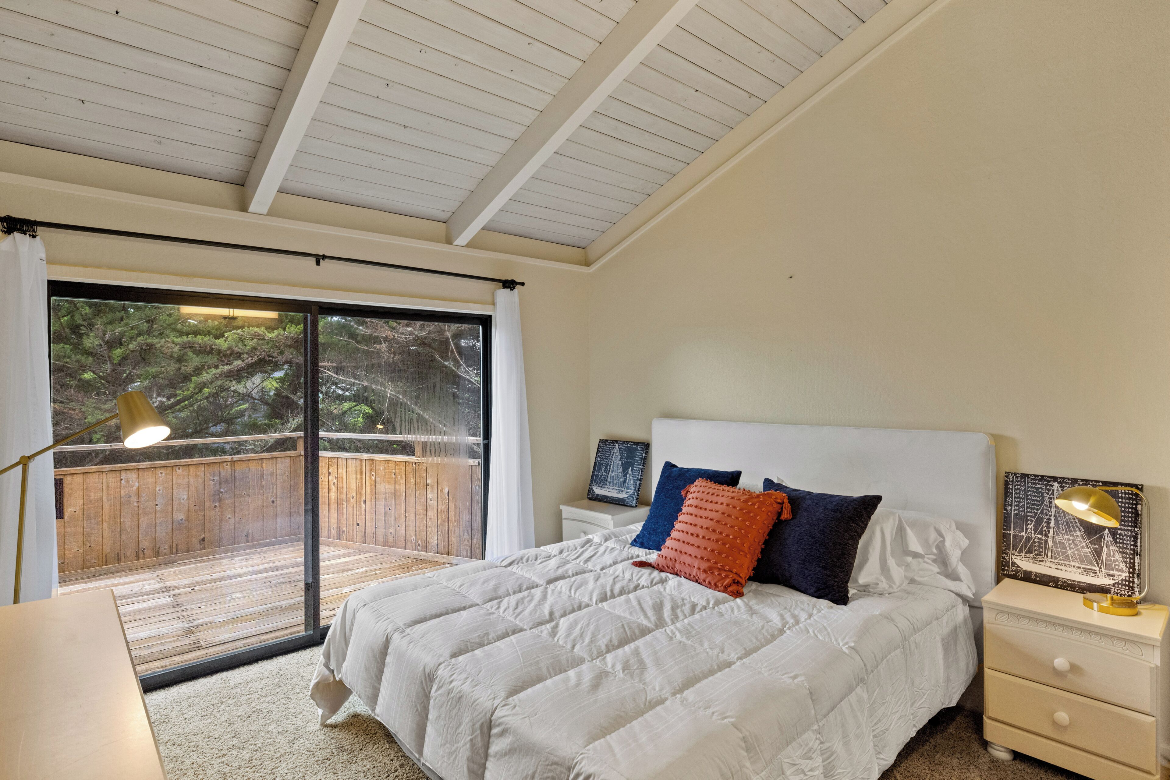 A bedroom with a large bed, a nightstand, a lamp, and a view of a wooden deck through a sliding glass door.
