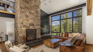 Ein modernes Wohnzimmer mit Kamin aus Stein, Ledersofa und großen Fenstern mit Blick auf einen Wald.