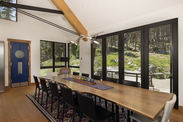 Un comedor con mesa de madera, sillas negras y vista a un bosque a través de ventanas amplias.