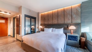 Een moderne hotelkamer met een groot bed, twee nachtkastjes met lampen, een inbouwkleerkast en uitzicht op de gang.