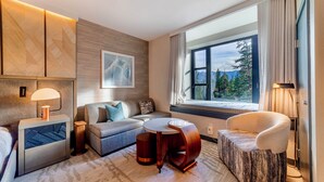 Een moderne hotelkamer met een bank, een ronde salontafel, een bijzettafeltje met een lamp, en uitzicht op een bos door een groot raam.