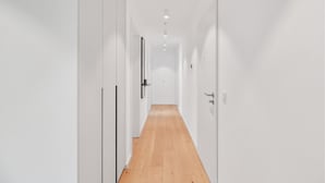 Un pasillo estrecho y bien iluminado, con paredes blancas y suelo de madera, que lleva a una puerta al final.