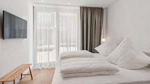 Un dormitorio moderno con cama, un banco, televisión y ventana con cortinas.