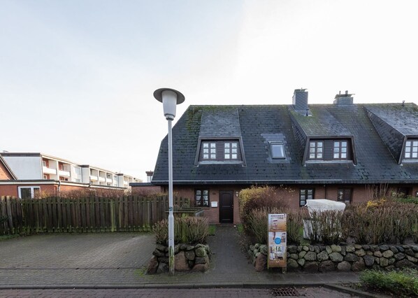 Ein zweistöckiges Gebäude mit geneigtem Dach, einem Hauseingang und einem kleinen Gartenbereich mit Steinmauer und einem Schild.