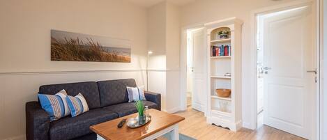 A living room with a dark grey sofa, a wooden coffee table, a blue rug, a bookshelf, and a white door.