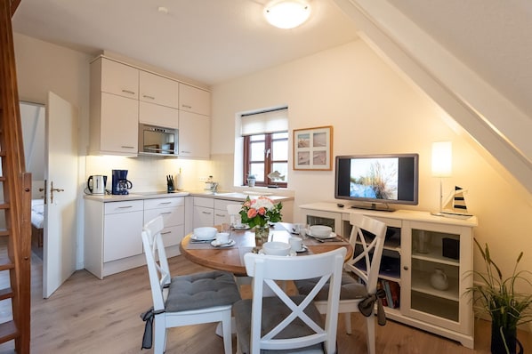 A modern kitchen with white cabinets, a dining table with chairs, a television, and a potted plant.