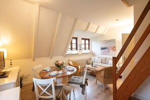 Ein Essbereich mit einem runden Tisch, gedeckt für vier Personen, umgeben von weißen Stühlen. Eine Rattan-Couch und ein kleiner Beistelltisch mit Blumen stehen dort. Im Vordergrund ist eine Holztreppe zu sehen.