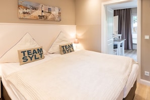 A hotel room with a large bed, two pillows with 'TEAM SYLT' text, a painting on the wall, and a desk with a chair in the background.