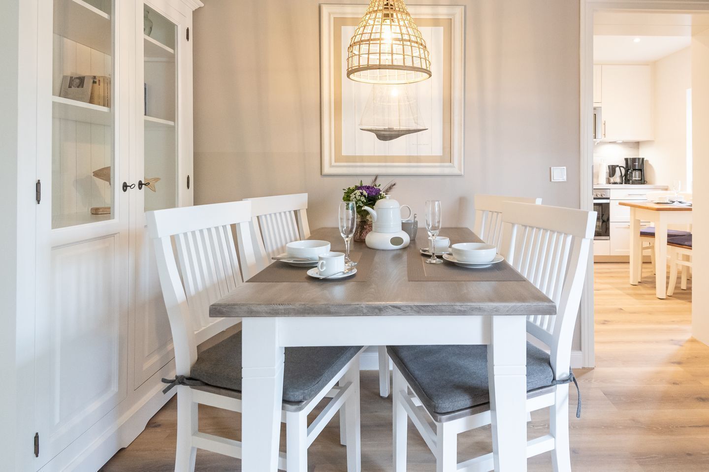 A dining area with a wooden table set for four, white chairs, a chandelier, and a kitchen in the background.