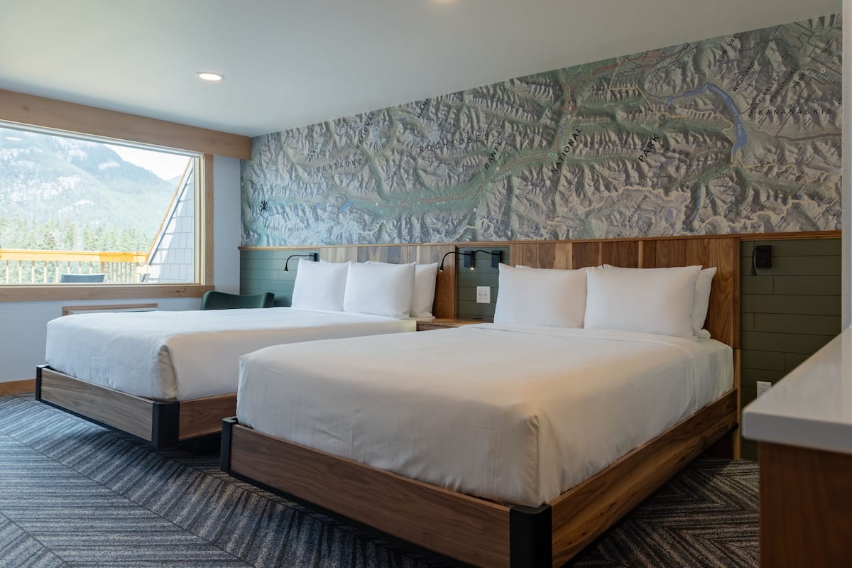 A hotel room with two beds, a large window with a mountain view, a map on the wall, and a wooden headboard.