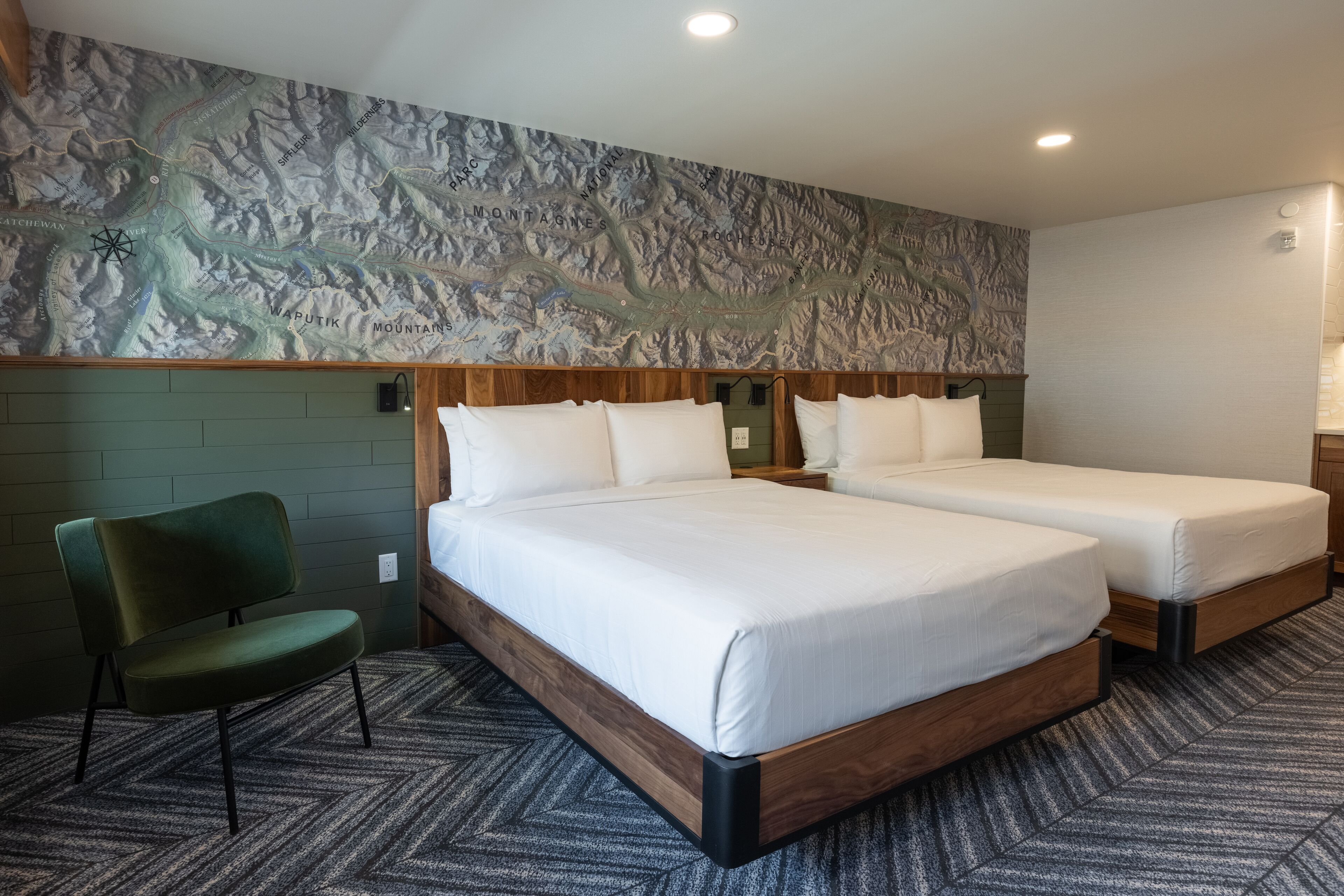 A hotel room with two beds, a green chair, and a mountain-themed wall.