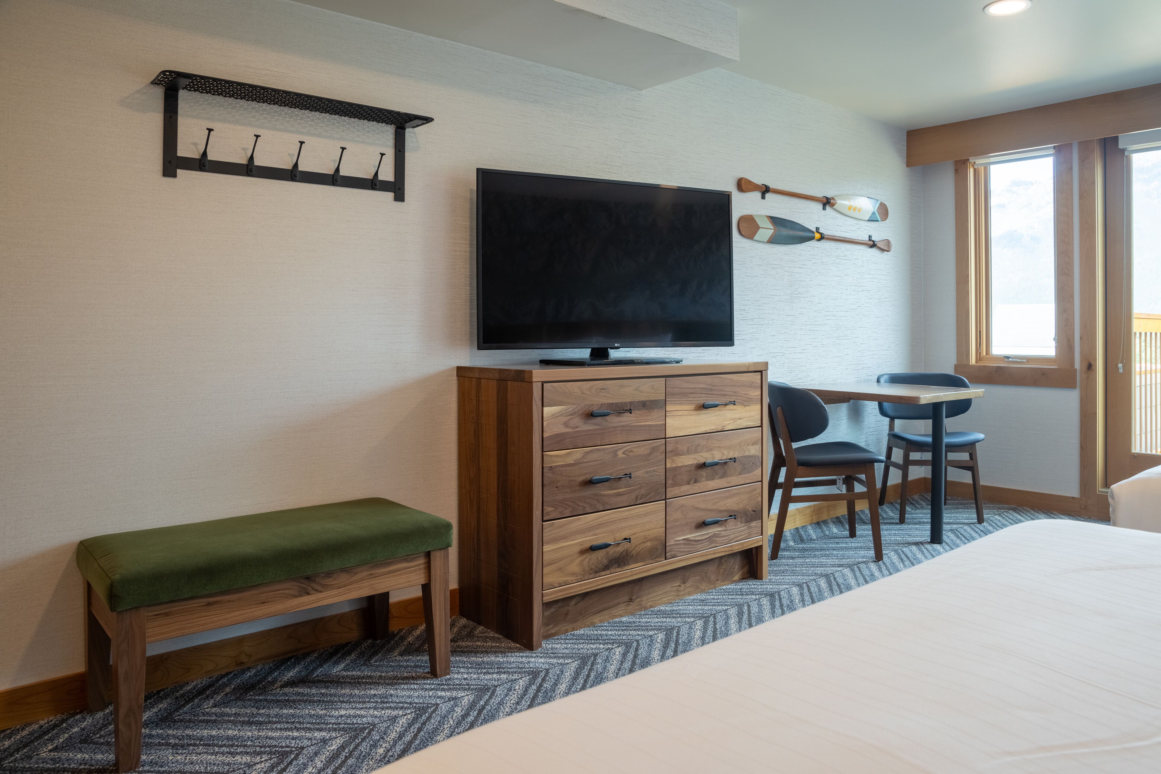 A hotel room with a bed, a wooden dresser, a television, a desk with two chairs, and a window.