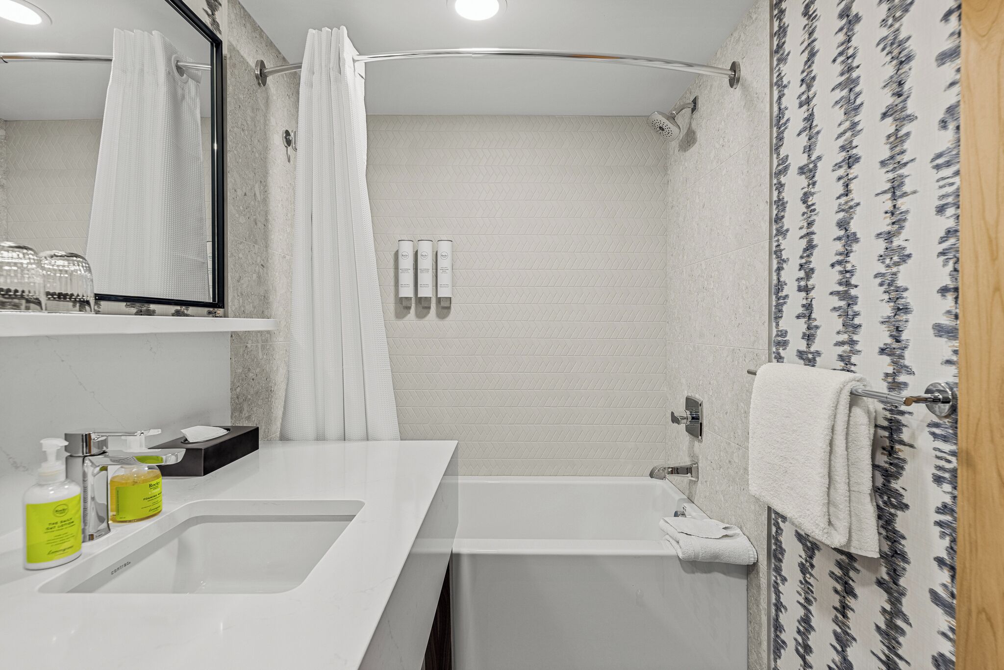 Un baño moderno con un lavabo blanco, espejo y zona de ducha. Hay artículos de aseo en el lavabo y un perchero con una toalla.