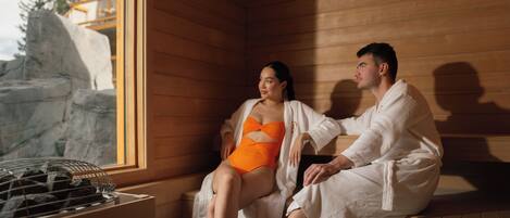 A couple in a wooden cabin with a sauna, a window, and a view of a rocky landscape.