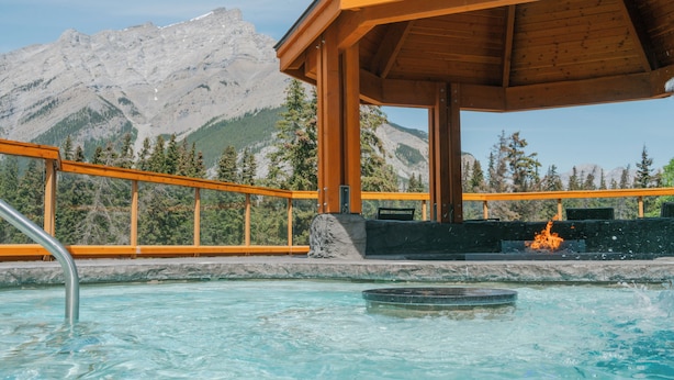 A wooden gazebo with a pool and a fire feature, surrounded by mountains and trees.